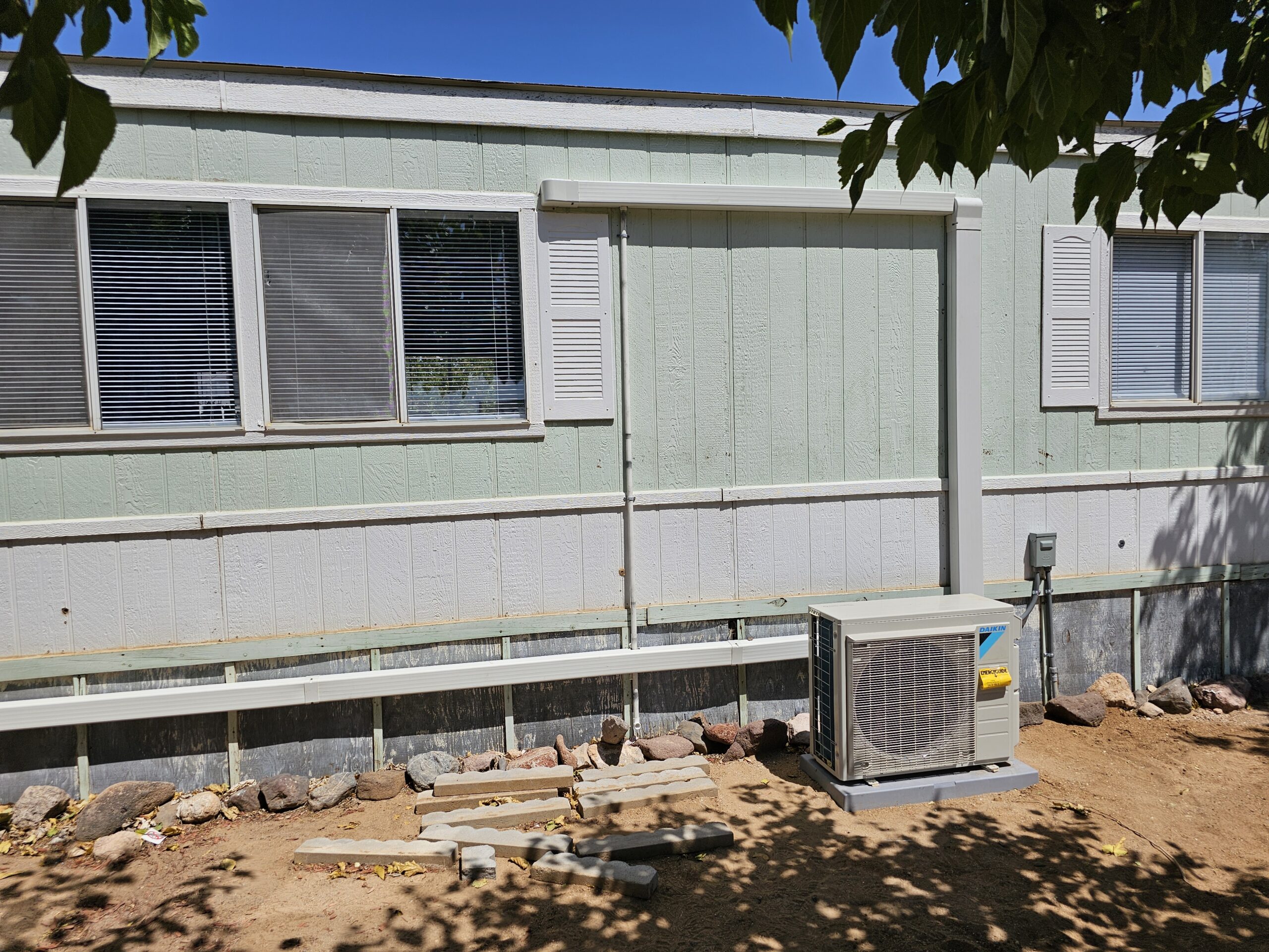 A light green mobile home with two windows, a white air conditioning unit installed outside, and rocks and dirt in the foreground.