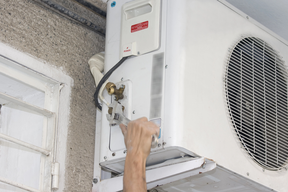 A person’s hand is adjusting a valve on an outdoor air conditioning unit mounted on a wall.