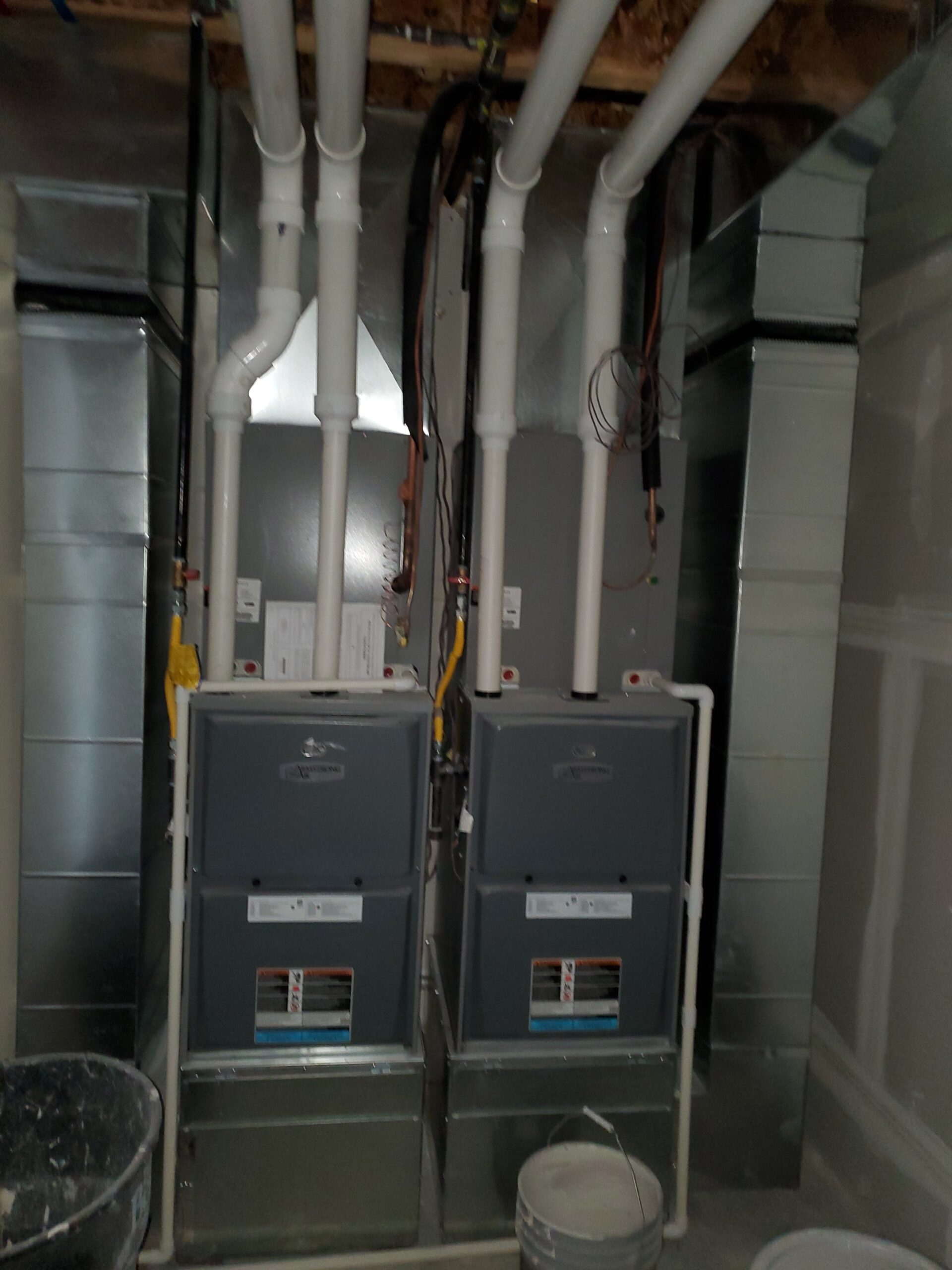 Two residential furnaces with multiple white PVC pipes and ductwork are installed side by side in a basement utility room.