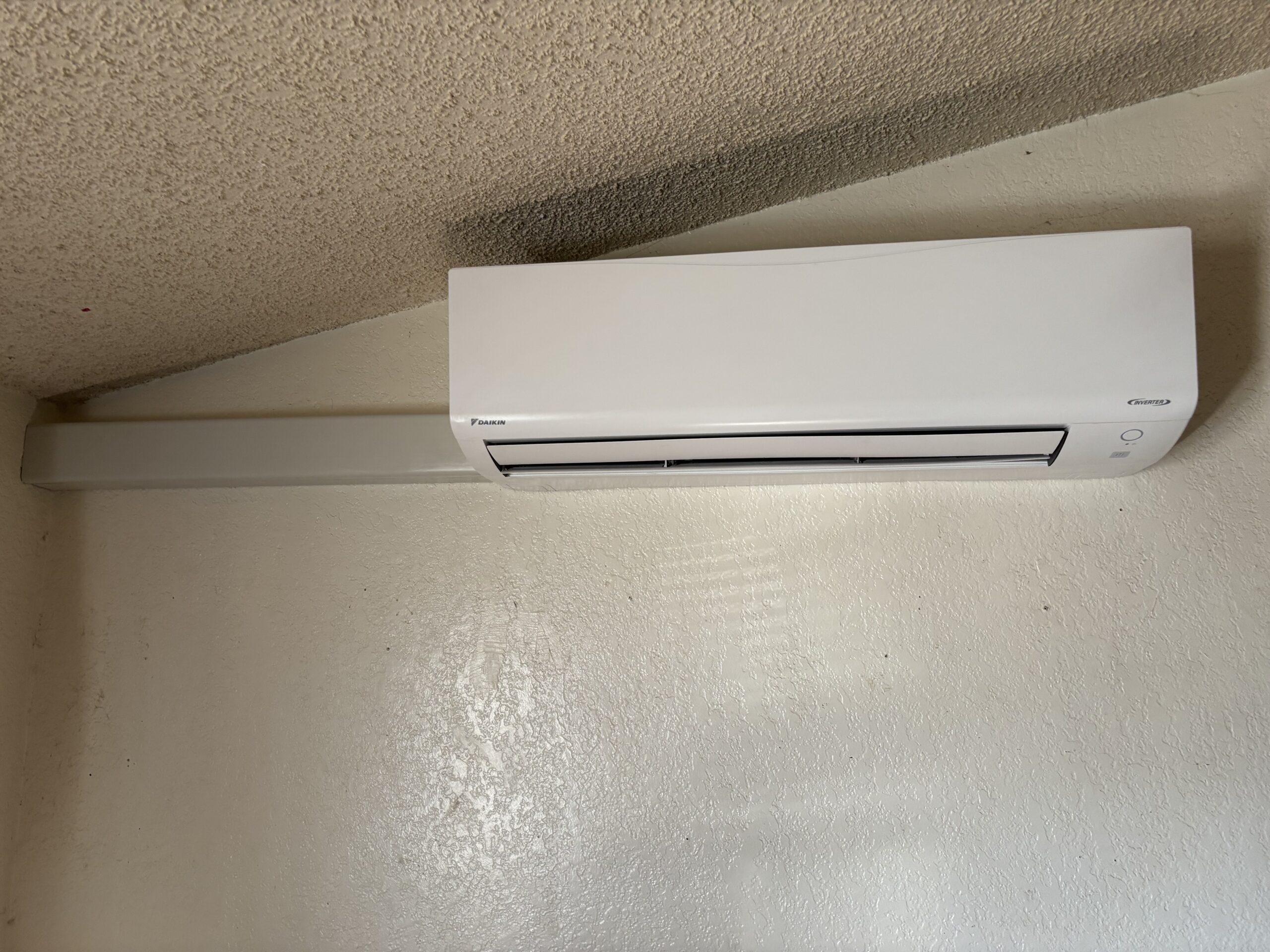 A white wall-mounted air conditioning unit with a horizontal cable cover extending to the left, installed near the ceiling on a textured wall.
